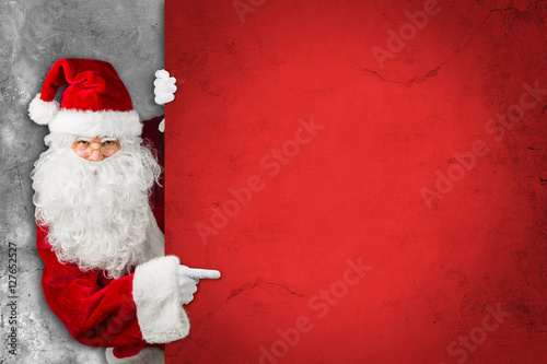 Santa claus hiding and looking through behind empty red concrete billboard wall and pointing with his finger on it