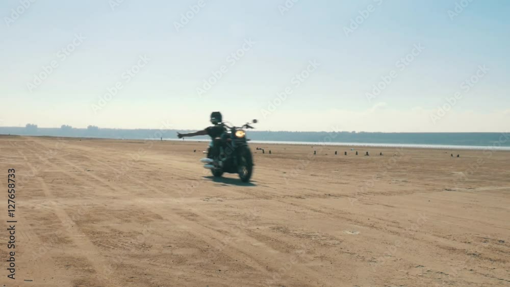 Motorcyclist riding along the sandy beach. Biker rides a motorcycle hands free through the desert. Chopper near the shore of the river, sea, lake, ocean. Front shot back light. Motorcyclist riding along the sandy beach. Biker rides a motorcycle hands free through the desert. Chopper near the shore of the river, sea, lake, ocean. Front shot back light.