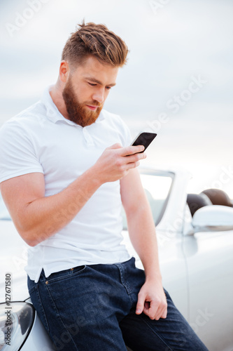 Concentrated bearded man with smart phone leaning on his car
