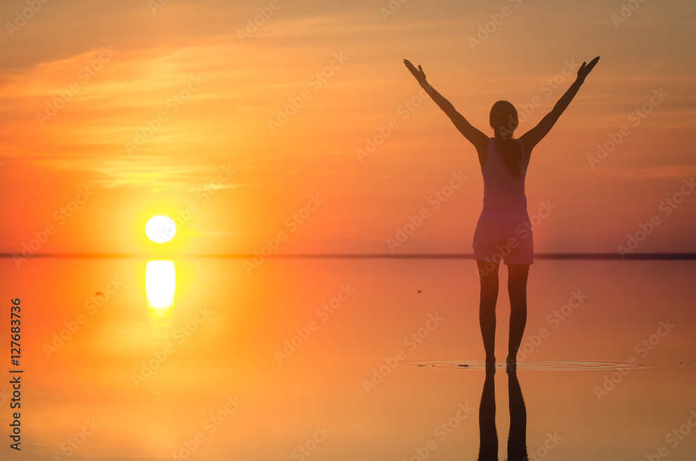 Beautiful female model open arms under sunrise at seaside. Calm water of salt lake Elton reflects woman silhouette. Sun goes behind horizon. Girl is alone