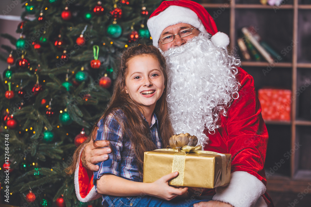 Christmas. Happy girl sitting at the hands of Santa. She holds a gift in the background Christmas tree Christmas. Happy girl sitting at the hands of Santa. She holds a gift in the background Christmas tree