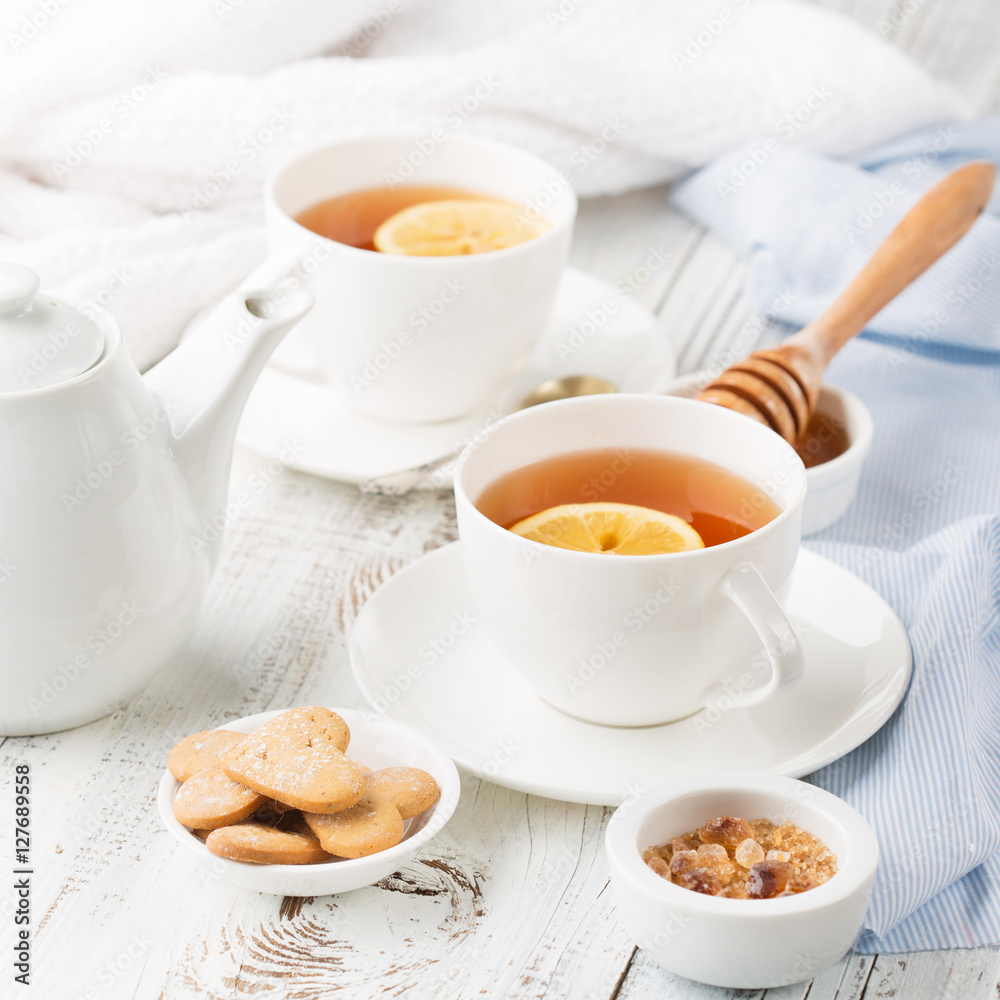 Two cups of hot black tea, lemon, homemade cookies and honey on white rustic wooden background. Selective focus Two cups of hot black tea, lemon, homemade cookies and honey on white rustic wooden background. Selective focus
