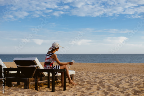 Back view of elegant lady relaxing on a chaise lounge, sandy beach outdoors background