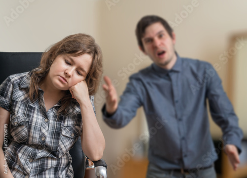 Young couple is arguing. Man is shouting and explaining something. Sad woman is sitting on chair and ignoring him.