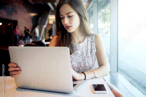 Young attractive woman is sitting in front of an open laptop in a coffee shop. Modern young business woman is working on a new project  looking at the screen of a portable computer.