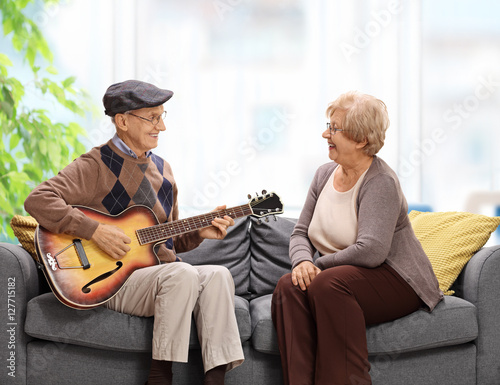 Elderly man sitting on a sofa and playing a guitar to an elderly