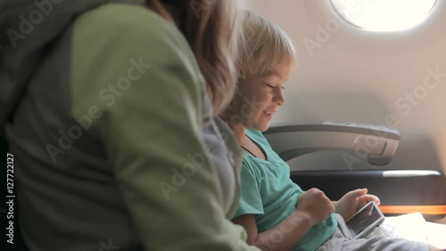 Cute little child sitting on his seat, playing games on smartphone, talking and smiling with mother traveling by plane. 