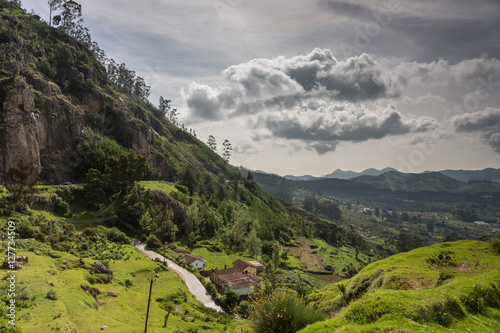 Ooty, India - October 25, 2013: Panoramic scenery of the Nilgiri hills shows forest, many hills at horizon, free floating stormy clouds, rocks, a road and a farm. Shades of green. Shot from us a hill. photo