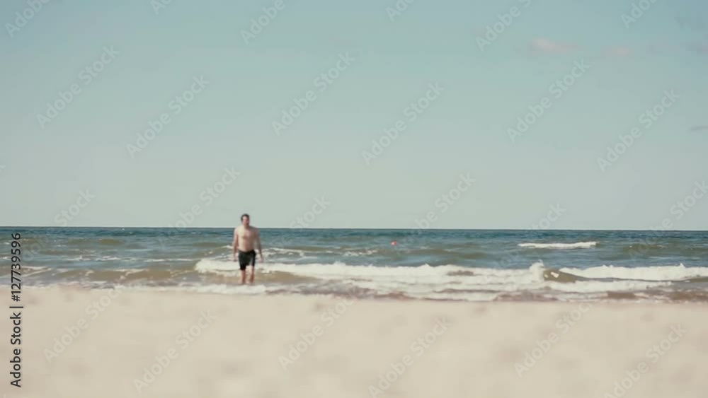 An athletic dark-haired young man wearing black swim shorts is coming out of the sea shirtless. An athletic dark-haired young man wearing black swim shorts is coming out of the sea shirtless.