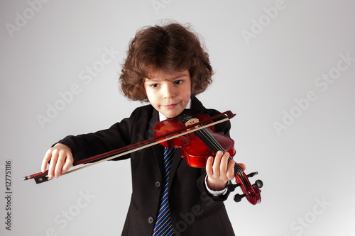 Small curly boy is playing a concert on the violin. Gray background.