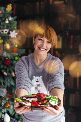 Portrait of beautiful redhead woman holding plate with christmas cakes before dressed up tree