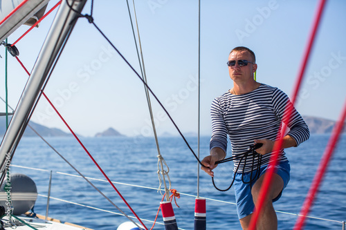 Young man skipper at his sail boat, controls ship during sea yacht race. Luxery vacations, sailing, adventure, travel.