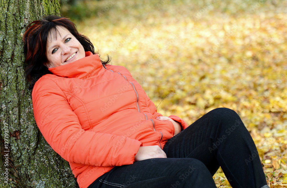 Obraz premium Portrait of smiling middle aged ordinary caucasian woman with long dark hair and red jacket leaning on tree.