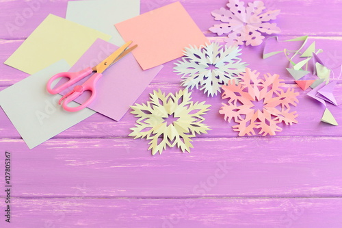 Colorful paper snowflakes, colored paper sheets and scrap, scissors on lilac wooden background. Cutting snowflakes out of colored paper. Simple winter crafts idea for children © onlynuta
