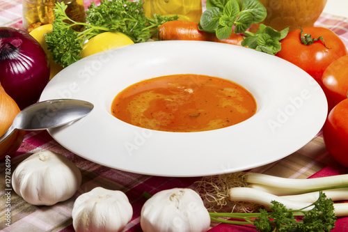 Red fish soup in a white plate beside fresh vegetables.