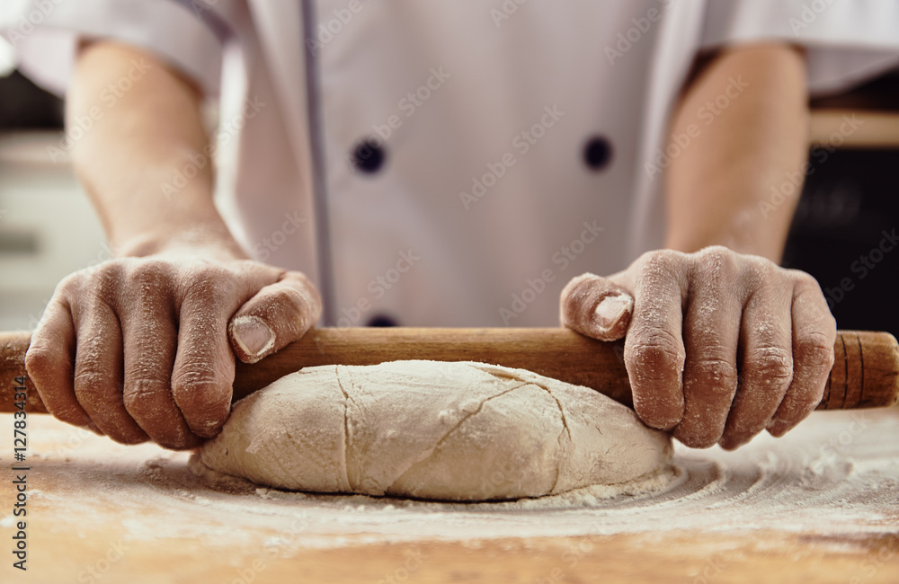 Cook's hands rolling raw dough with pin in rustic kitchen. 