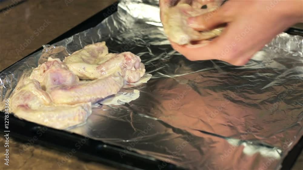 Young woman puts chicken wings on baking sheet with foil. Woman in kitchen preparing food. Housewife engaged in household chores in kitchen Young woman puts chicken wings on baking sheet with foil. Woman in kitchen preparing food. Housewife engaged in household chores in kitchen