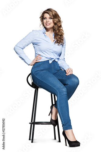 Beautiful fashionable plump woman in jeans and a denim shirt sits on a chair on a white background