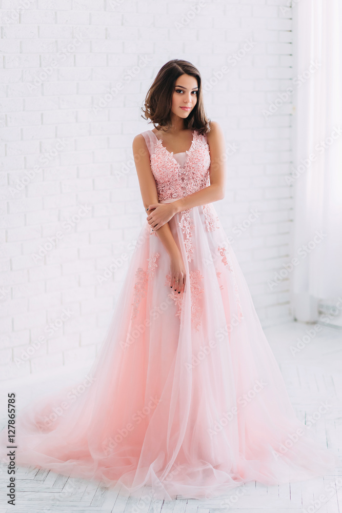 Beautiful brunette girl standing in a bright room. She is dressed in luxurious, fluffy, pink dress. Gentle puppet's face looking at the camera. The European image of the bride. Photo high key style. Beautiful brunette girl standing in a bright room. She is dressed in luxurious, fluffy, pink dress. Gentle puppet's face looking at the camera. The European image of the bride. Photo high key style.