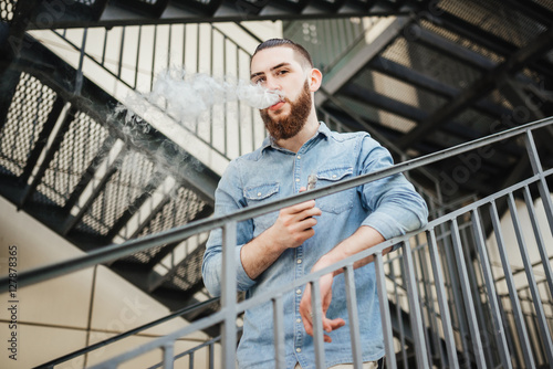 Young man with beard vaping an electronic cigarette outdoor. Casual hipster smoke vaporizer. Vaping