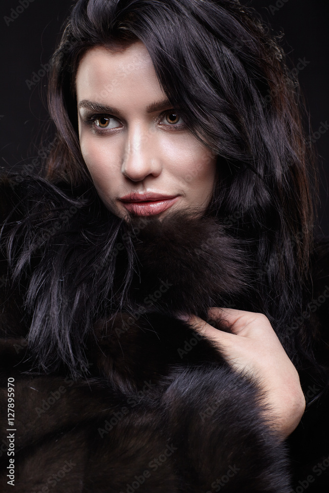 Portrait of a young brunette woman with long black hair dressed in a fur coat on black background Portrait of a young brunette woman with long black hair dressed in a fur coat on black background