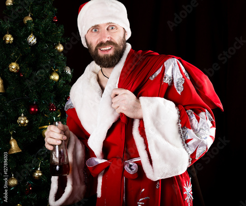 bad brutal Santa Claus carries a bag, smiling spitefully and drinking brandy from a bottle, on the background of Christmas tree photo
