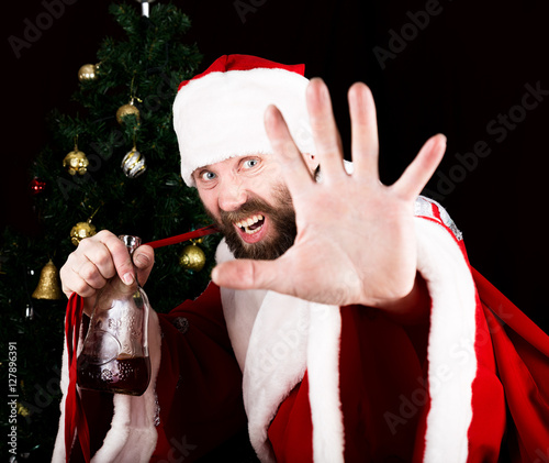 bad brutal Santa Claus smiling spitefully, drinking brandy from a bottle and outstretched arm, on the background of Christmas tree photo