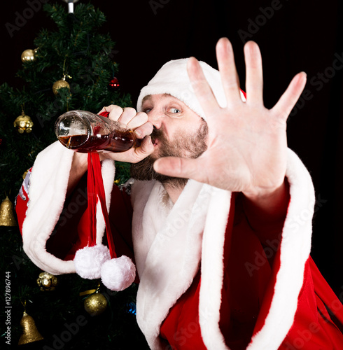 bad brutal Santa Claus smiling spitefully, drinking brandy from a bottle and outstretched arm, on the background of Christmas tree photo