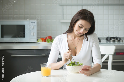 Portrait of happy attractive woman sitting at the kitchen table  having healthy lunch  salad and orange juice  holding smartphone