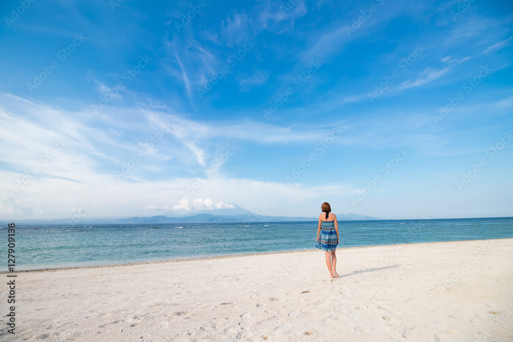 Young woman in blue dress walking on beach of tropical island Nusa Lembongan, Indonesia. Amazing sky, ocean view. Volcano Agung and island Bali on the background. Young woman in blue dress walking on beach of tropical island Nusa Lembongan, Indonesia. Amazing sky, ocean view. Volcano Agung and island Bali on the background.