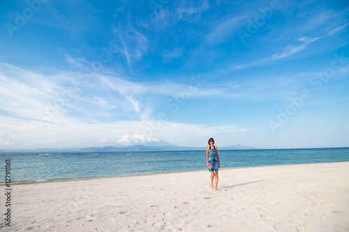Young woman in blue dress walking on beach of tropical island Nusa Lembongan, Indonesia. Amazing sky, ocean view. Volcano Agung and island Bali on the background.