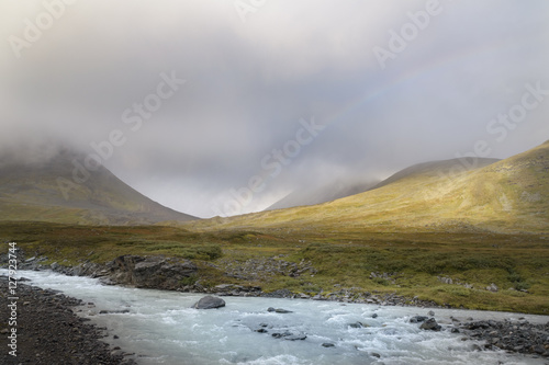 Stream of melted water flowing through tranquil landscape with rainbow between the mountain background photo