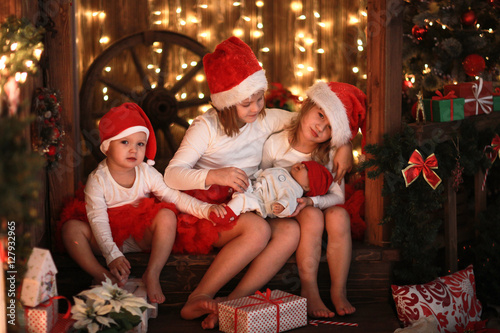 Siblings sisters in santa hats with doll near fireplace on Chri