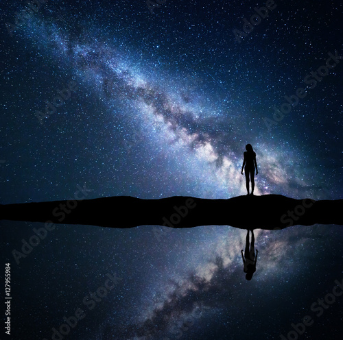 Milky Way. Night starry sky and silhouette of a standing woman on the mountain near the lake with sky reflection in water. Landscape with blue Milky Way and woman. Galaxy, Universe. Space background