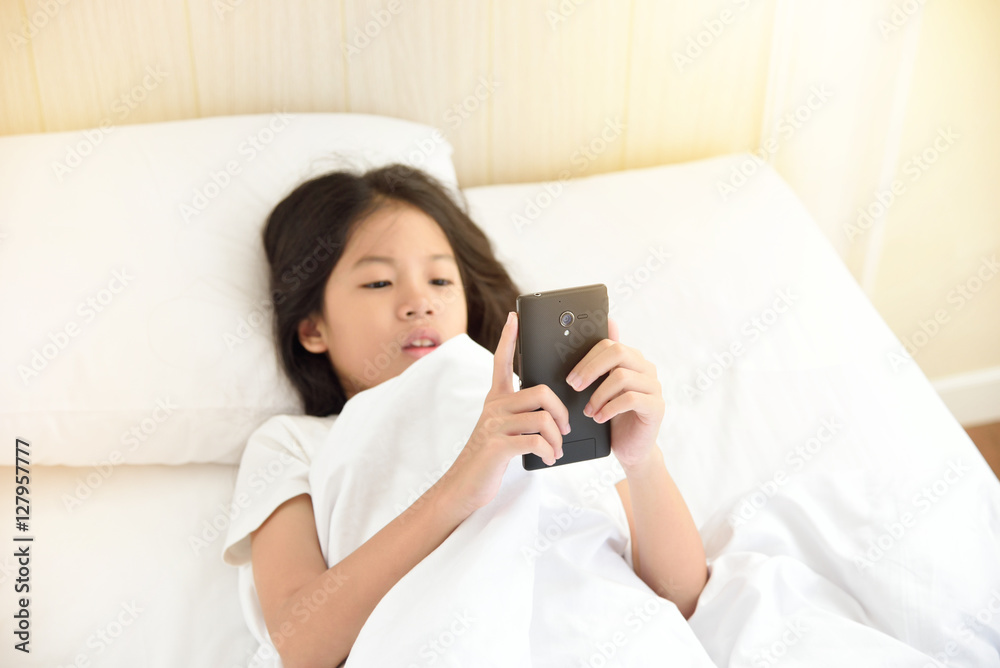 Concentrated young Asian girl looking at her smartphone lying on the bed in bedroom. Technology and internet concept. Copy space. Preteen or school age girl using smart phone on the bed in the morning Concentrated young Asian girl looking at her smartphone lying on the bed in bedroom. Technology and internet concept. Copy space. Preteen or school age girl using smart phone on the bed in the morning
