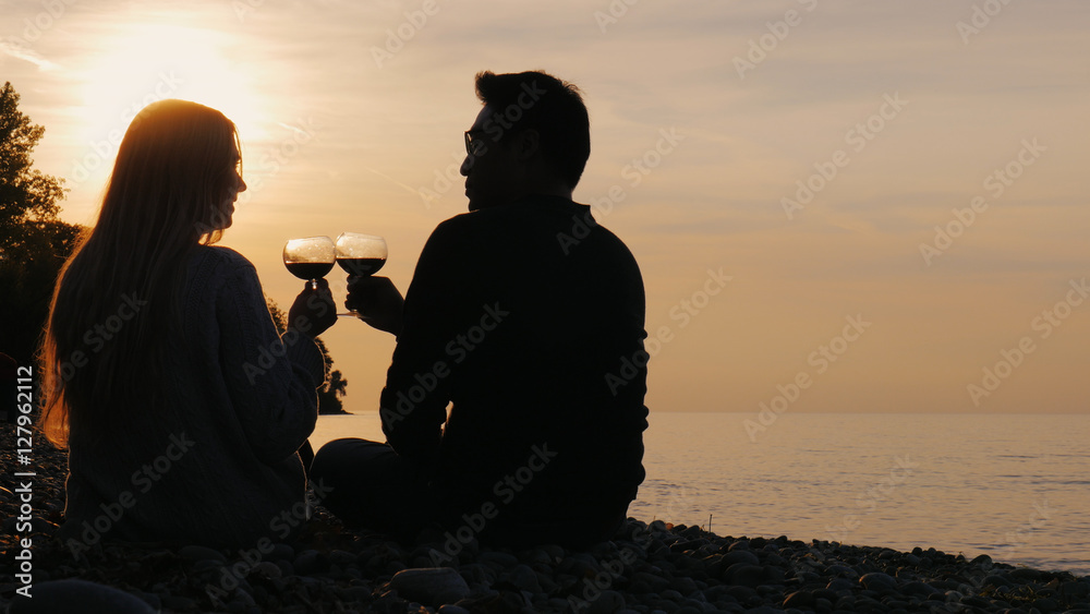 Silhouettes of a young Asian man and Caucasian woman. Sit on the beach at sunset, drink wine from glasses

