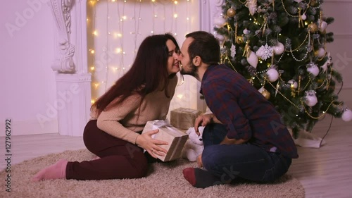 A young man gives his girlfriend for Christmas. The lovers sit in front of the fireplace on the beige carpet.