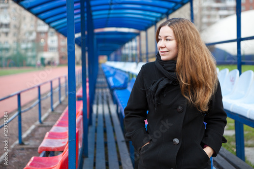 Young woman in a black coat sitting on an empty grandstand outdoor stadium and looking away