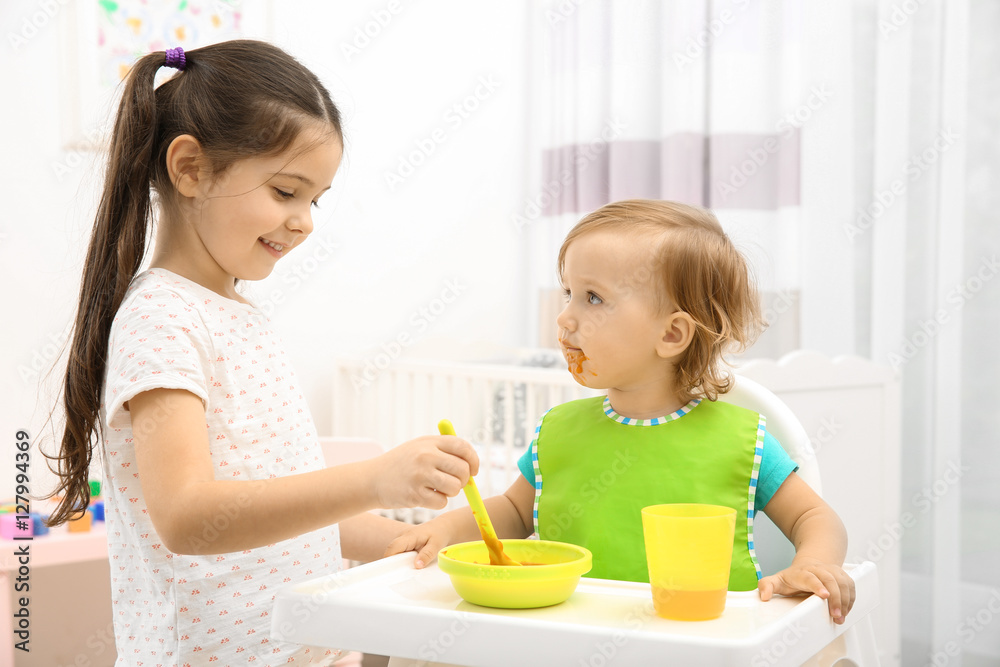 Cute girl feeding little sister in high chair at baby room