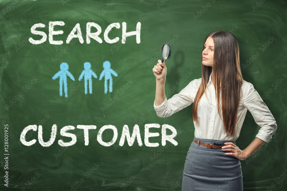 Beautiful businesswoman standing and looking through the magnifying glass near the green wall with words 'search customer' drawn on it.