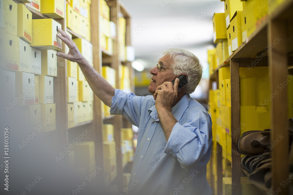 Senior man on the phone in warehouse with shoe boxes