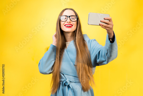 Young and smiling woman in the blue coat making self portrait with smart phone standing on the yellow wall background