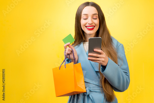 Stylish woman with shopping bag, credit card and phone on the yellow wall background. Winter holiday sale