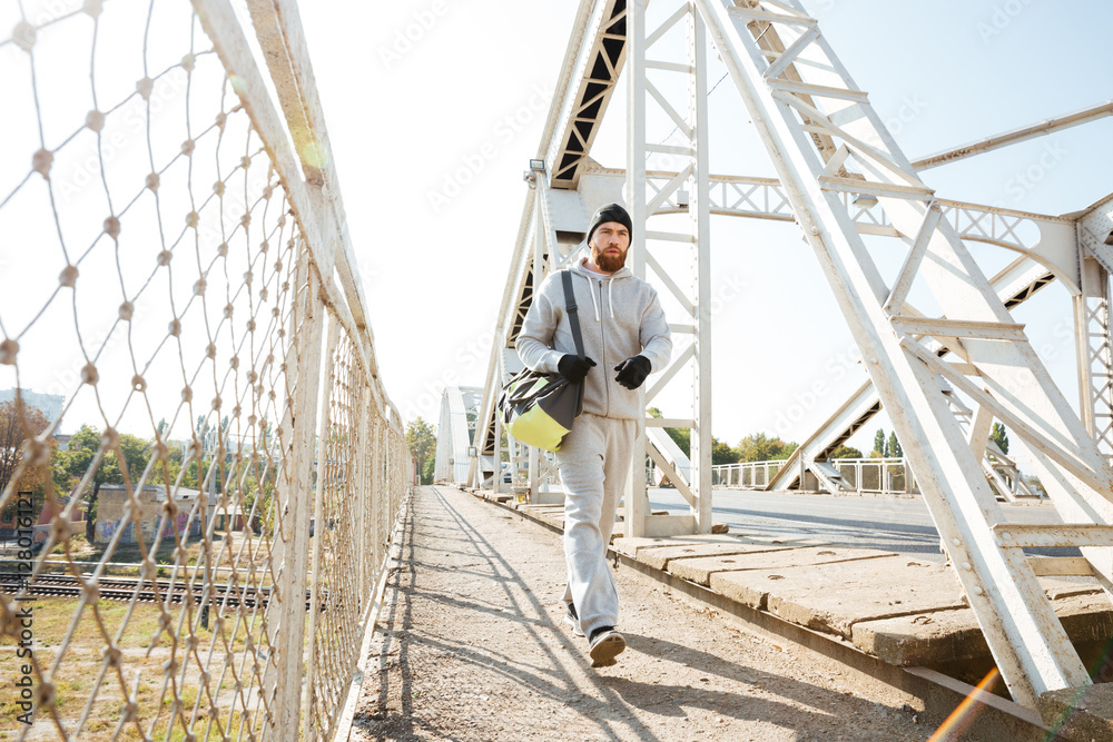 Young bearded sports man with bag walking along urban bridge