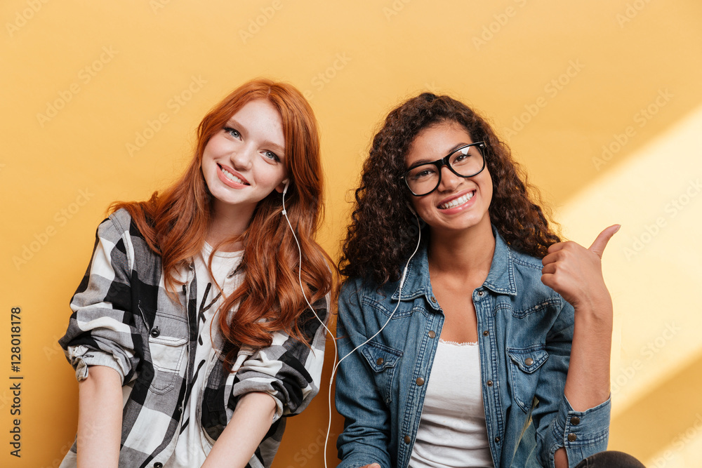 Fototapeta premium Two happy lovely young women with earphones showing thumbs up