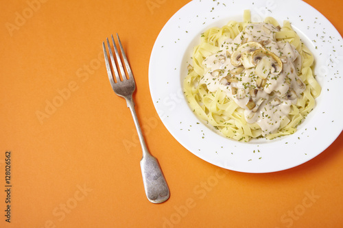 A dinner dish full of tagliatelle spaghetti with a creamy mushroom pasta sauce, on a bright orange background with fork and blank space at side