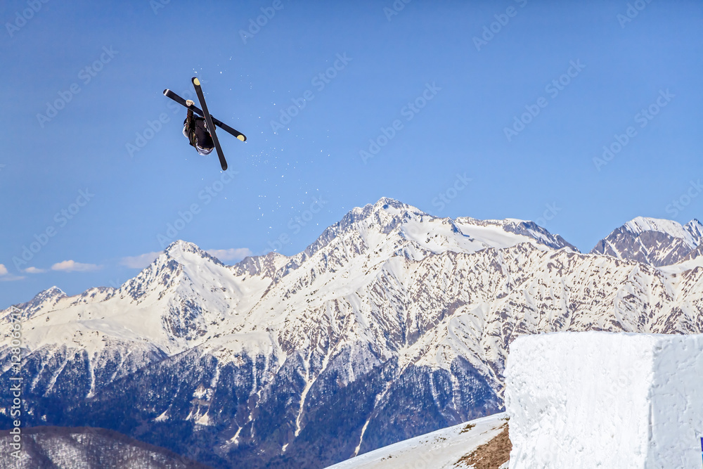 Skier flying from a ski jump making figure on blue sky and snowy mountain peaks background Skier flying from a ski jump making figure on blue sky and snowy mountain peaks background