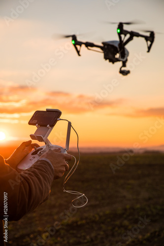 Man controls the fly of quadrocopter in field over sunset background