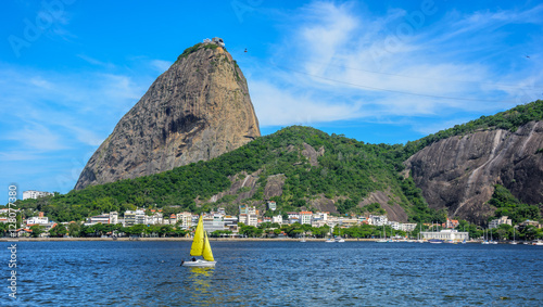 Yellow small sailing yacht and classic daytime scenic profile view of Sugarloaf Mountain, Pao de Acucar, standing above Botafogo Bay in Rio de Janeiro, Brazil