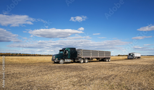 Two semi trucks with attached grain trailers parked in a harvested field under cloudy sky in rural autumn landscape
